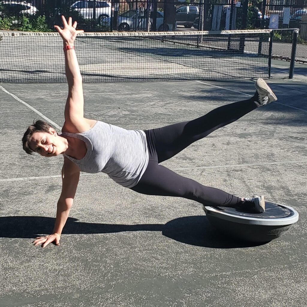 Woman in gray shirt doing a side plank with top leg raised and bottom leg on a bosu ball | virtual fitness program